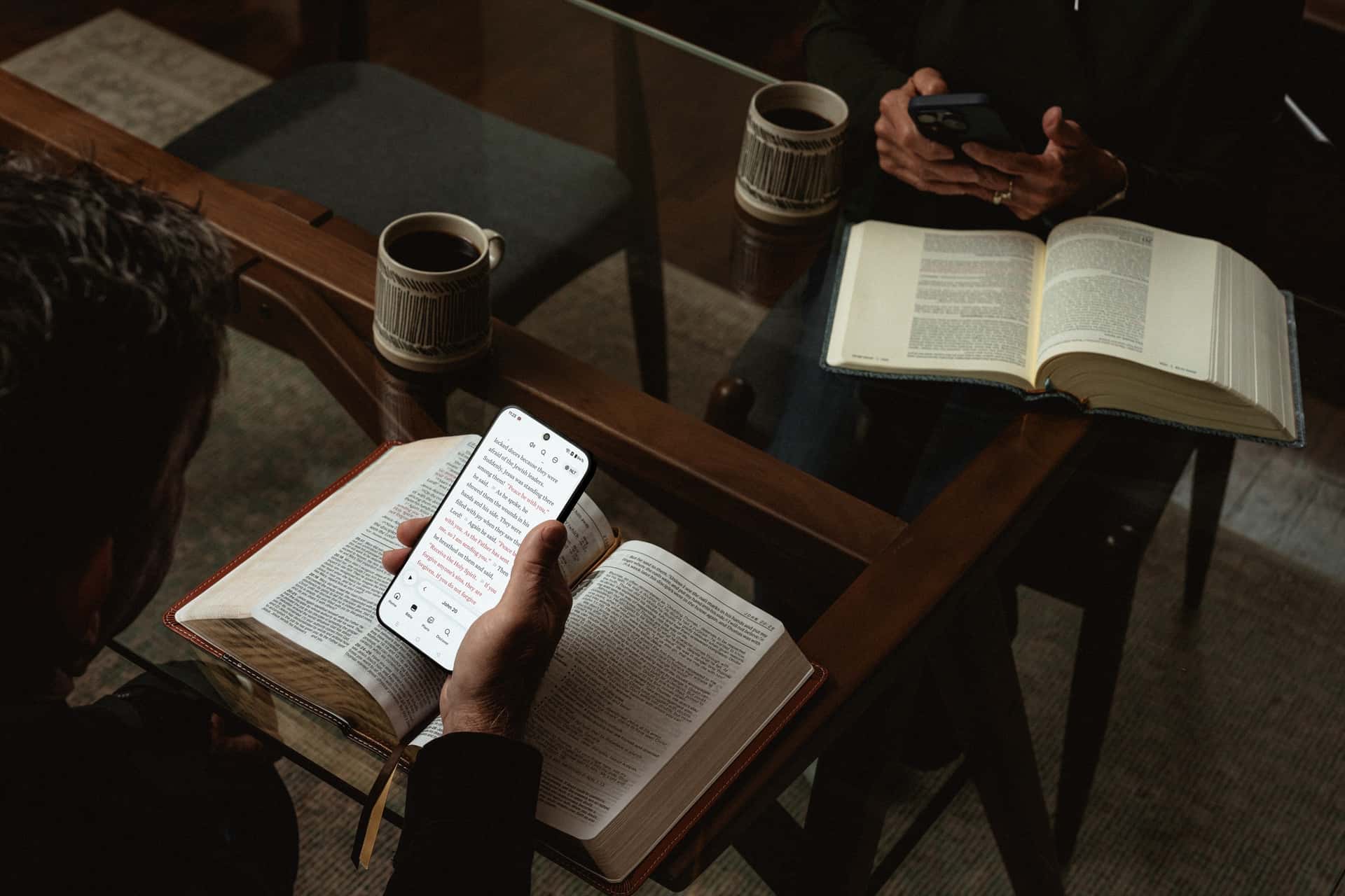 Two people reading open Bibles at a wooden table, one following along on a phone, with coffee beside them.